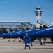 A row of U.S. Navy Blue Angels F/A-18 Hornet jets are parked on an airport tarmac, identifiable by their distinctive blue and gold paint scheme. Personnel in blue flight suits are walking near and climbing on the aircraft. A large airport control tower and terminal building are visible in the background, suggesting this is a major civilian or joint-use airport.