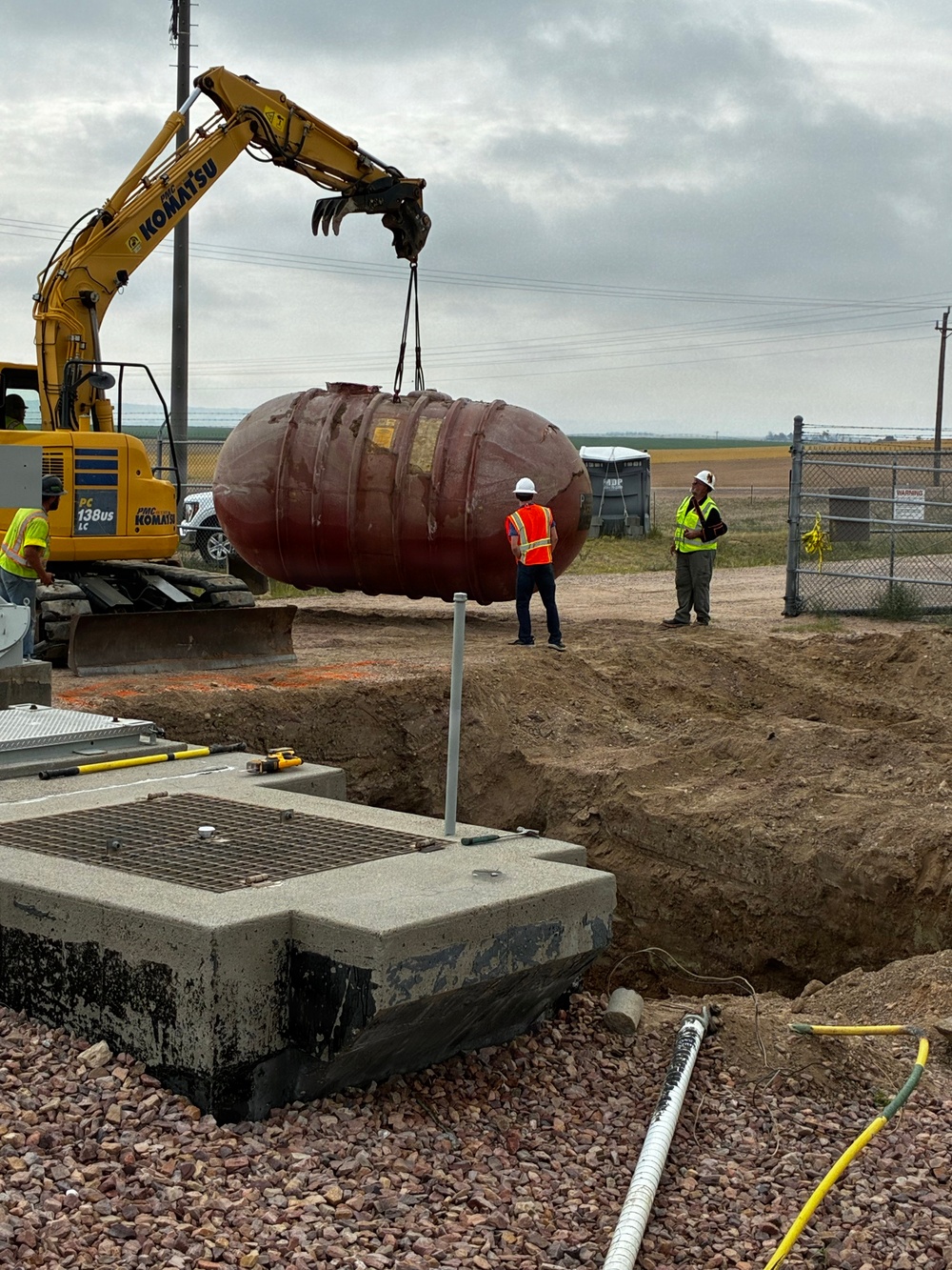 A Komatsu PC138US-LC excavator is lifting a large oval/elliptical underground storage tank (UST) using chains at what appears to be a fuel station or industrial construction site. The tank, which appears to be a fiberglass or steel UST with a dark reddish-brown exterior, is being removed from or installed into an excavated pit. Three workers in high-visibility safety gear and hard hats are present, overseeing the operation.