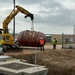 A Komatsu PC138US-LC excavator is lifting a large oval/elliptical underground storage tank (UST) using chains at what appears to be a fuel station or industrial construction site. The tank, which appears to be a fiberglass or steel UST with a dark reddish-brown exterior, is being removed from or installed into an excavated pit. Three workers in high-visibility safety gear and hard hats are present, overseeing the operation.