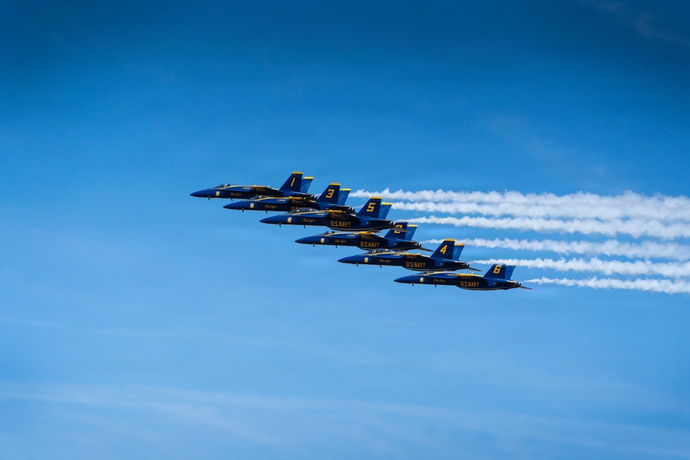 Six U.S. Navy Blue Angels aircraft are flying in a tight diagonal echelon formation against a clear blue sky. The jets, numbered 1 through 6, are producing white smoke trails as they perform a formation flight display. The aircraft are painted in the iconic blue and gold color scheme with 'U.S. NAVY' markings clearly visible.