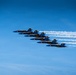 Six U.S. Navy Blue Angels aircraft are flying in a tight diagonal echelon formation against a clear blue sky. The jets, numbered 1 through 6, are producing white smoke trails as they perform a formation flight display. The aircraft are painted in the iconic blue and gold color scheme with 'U.S. NAVY' markings clearly visible.