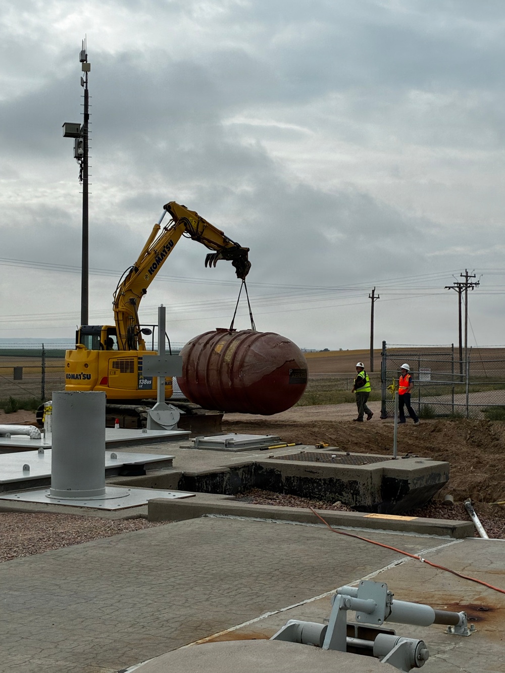 A Komatsu excavator is lifting a large red/maroon elliptical tank or vessel at what appears to be an industrial or utility construction site. Two workers wearing hard hats and high-visibility vests are observing the lift operation nearby. The site features concrete pads, underground vault structures, and various mechanical components in a rural, flat landscape.