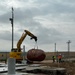 A Komatsu excavator is lifting a large red/maroon elliptical tank or vessel at what appears to be an industrial or utility construction site. Two workers wearing hard hats and high-visibility vests are observing the lift operation nearby. The site features concrete pads, underground vault structures, and various mechanical components in a rural, flat landscape.