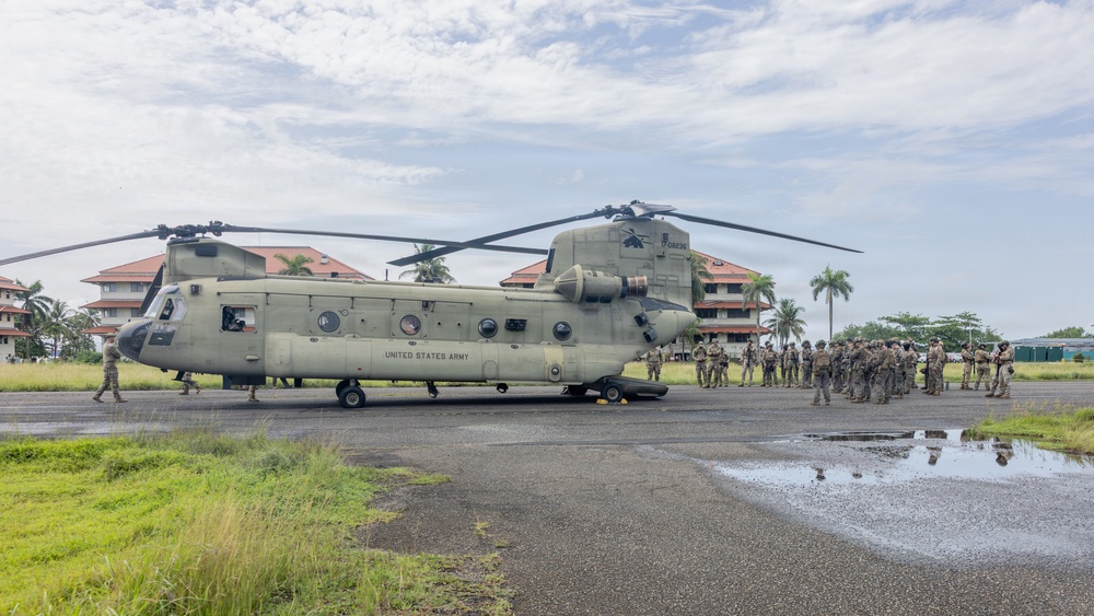 A United States Army CH-47 Chinook tandem-rotor helicopter is parked on a wet tarmac in what appears to be a tropical location. A large group of military personnel in combat gear are assembled near the rear of the aircraft, likely receiving a briefing or preparing to board. The setting includes palm trees and red-roofed buildings in the background, suggesting a tropical or Southeast Asian environment.