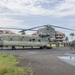 A United States Army CH-47 Chinook tandem-rotor helicopter is parked on a wet tarmac in what appears to be a tropical location. A large group of military personnel in combat gear are assembled near the rear of the aircraft, likely receiving a briefing or preparing to board. The setting includes palm trees and red-roofed buildings in the background, suggesting a tropical or Southeast Asian environment.
