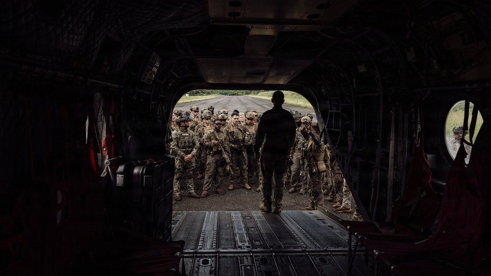 The image is taken from inside the rear cargo bay of a large military helicopter, likely a CH-47 Chinook, looking outward through the open ramp. A silhouetted figure stands at the ramp's edge facing a large group of fully equipped soldiers assembled on a tarmac or airstrip. The scene suggests a pre-mission briefing or troops preparing to board the aircraft.