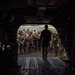 The image is taken from inside the rear cargo bay of a large military helicopter, likely a CH-47 Chinook, looking outward through the open ramp. A silhouetted figure stands at the ramp's edge facing a large group of fully equipped soldiers assembled on a tarmac or airstrip. The scene suggests a pre-mission briefing or troops preparing to board the aircraft.