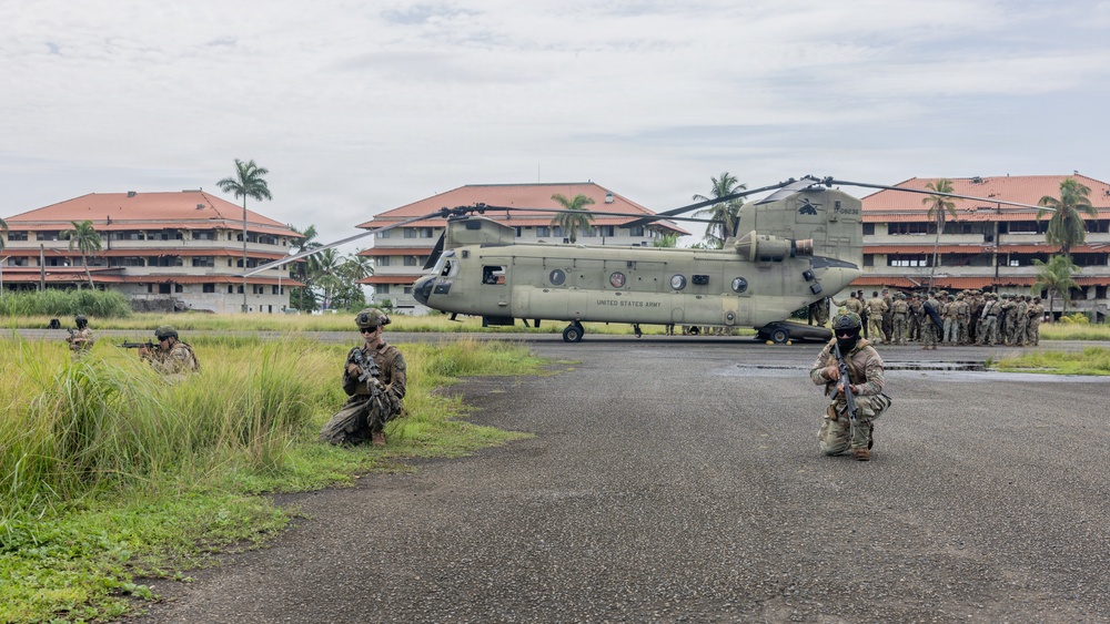 A United States Army CH-47 Chinook helicopter is landed on a tarmac or airstrip in what appears to be a tropical location, based on the palm trees and red-roofed buildings in the background. Several soldiers in full combat gear are positioned in defensive postures around the aircraft, while a larger group of troops is assembled to the right. The scene appears to depict a military training exercise or tactical insertion operation.