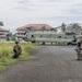 A United States Army CH-47 Chinook helicopter is landed on a tarmac or airstrip in what appears to be a tropical location, based on the palm trees and red-roofed buildings in the background. Several soldiers in full combat gear are positioned in defensive postures around the aircraft, while a larger group of troops is assembled to the right. The scene appears to depict a military training exercise or tactical insertion operation.