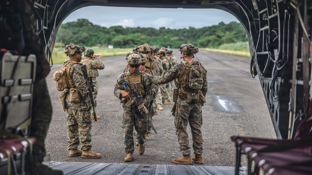 Military personnel in full combat gear are boarding or staging near the open rear ramp of a large military helicopter, likely a CH-47 Chinook, on a tarmac or airstrip. The soldiers are wearing woodland MARPAT camouflage uniforms consistent with U.S. Marines, equipped with tactical vests, helmets, and rifles. The setting appears to be a tropical or subtropical environment based on the lush green vegetation visible in the background.