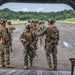 Military personnel in full combat gear are boarding or staging near the open rear ramp of a large military helicopter, likely a CH-47 Chinook, on a tarmac or airstrip. The soldiers are wearing woodland MARPAT camouflage uniforms consistent with U.S. Marines, equipped with tactical vests, helmets, and rifles. The setting appears to be a tropical or subtropical environment based on the lush green vegetation visible in the background.