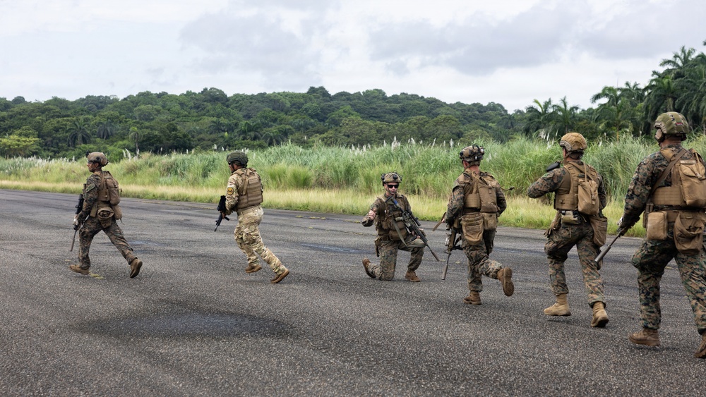 A group of armed military personnel in full combat gear are moving across what appears to be a tarmac or airstrip in a tropical environment. The soldiers are wearing mixed camouflage patterns, suggesting possible multinational or multi-branch composition. The setting features dense tropical vegetation in the background, consistent with a jungle or equatorial region.