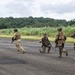 A group of armed military personnel in full combat gear are moving across what appears to be a tarmac or airstrip in a tropical environment. The soldiers are wearing mixed camouflage patterns, suggesting possible multinational or multi-branch composition. The setting features dense tropical vegetation in the background, consistent with a jungle or equatorial region.