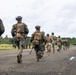 A group of heavily equipped military personnel in combat gear are running along a paved airstrip or road in what appears to be a tropical environment. The soldiers are wearing a mix of camouflage uniforms, tactical vests, helmets with mounted equipment, and carrying backpacks and weapons. The scene suggests a training exercise or tactical movement operation.