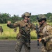 Two heavily equipped military personnel in different camouflage patterns are engaged in what appears to be a briefing or coordination on a tarmac or paved surface. The foreground soldier, wearing MARPAT (Marine pattern) camouflage with advanced helmet-mounted optics, is gesturing directionally toward another soldier in multicam pattern. Additional military personnel are visible in the background on a grassy field bordered by tropical vegetation.