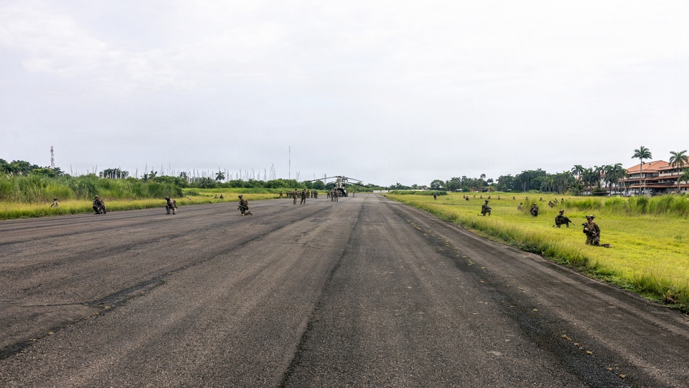 Military personnel are deployed along both sides of a runway or airstrip in a tropical environment, with a large helicopter (appears to be a CH-47 Chinook or similar heavy-lift helicopter) visible in the distance at the far end of the strip. Soldiers are positioned in kneeling and prone security postures on and beside the tarmac, suggesting a tactical landing zone security operation or military exercise. The surrounding vegetation, palm trees, and architecture suggest a tropical or subtropical location.