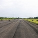 Military personnel are deployed along both sides of a runway or airstrip in a tropical environment, with a large helicopter (appears to be a CH-47 Chinook or similar heavy-lift helicopter) visible in the distance at the far end of the strip. Soldiers are positioned in kneeling and prone security postures on and beside the tarmac, suggesting a tactical landing zone security operation or military exercise. The surrounding vegetation, palm trees, and architecture suggest a tropical or subtropical location.