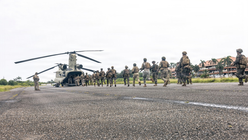 A column of military personnel in full combat gear is boarding a CH-47 Chinook tandem-rotor helicopter on a tarmac or airstrip. The soldiers are equipped with tactical vests, helmets, and weapons, moving in an organized line toward the rear ramp of the aircraft. The setting appears to be a tropical location, evidenced by palm trees and lush vegetation visible in the background.