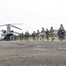 A column of military personnel in full combat gear is boarding a CH-47 Chinook tandem-rotor helicopter on a tarmac or airstrip. The soldiers are equipped with tactical vests, helmets, and weapons, moving in an organized line toward the rear ramp of the aircraft. The setting appears to be a tropical location, evidenced by palm trees and lush vegetation visible in the background.