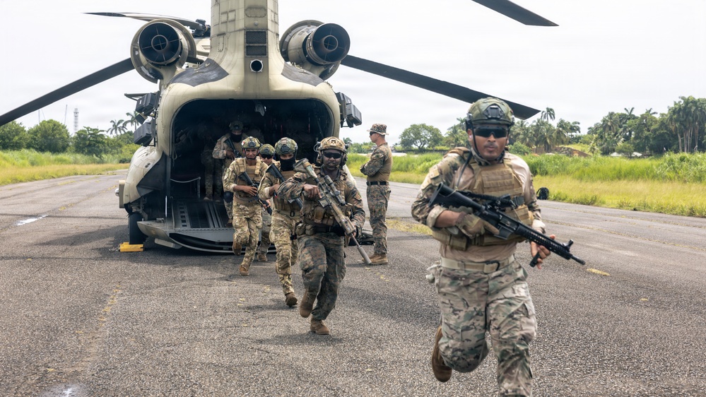 Military personnel in full combat gear are rapidly disembarking from a CH-47 Chinook tandem-rotor helicopter onto a tarmac or airstrip. The soldiers are armed with assault rifles and wearing camouflage uniforms and tactical helmets. The setting appears to be a tropical environment based on the lush green vegetation and palm trees visible in the background.