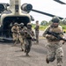 Military personnel in full combat gear are rapidly disembarking from a CH-47 Chinook tandem-rotor helicopter onto a tarmac or airstrip. The soldiers are armed with assault rifles and wearing camouflage uniforms and tactical helmets. The setting appears to be a tropical environment based on the lush green vegetation and palm trees visible in the background.