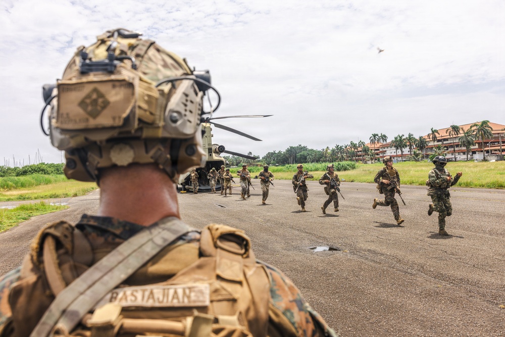Military personnel are conducting a helicopter insertion/extraction exercise on a tarmac or airstrip in a tropical environment. Soldiers are running away from a CH-47 Chinook helicopter in the background while a Marine identified as 'Bastajian' observes from the foreground. The setting appears to be a joint military exercise given the mix of camouflage patterns visible.