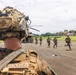 Military personnel are conducting a helicopter insertion/extraction exercise on a tarmac or airstrip in a tropical environment. Soldiers are running away from a CH-47 Chinook helicopter in the background while a Marine identified as 'Bastajian' observes from the foreground. The setting appears to be a joint military exercise given the mix of camouflage patterns visible.