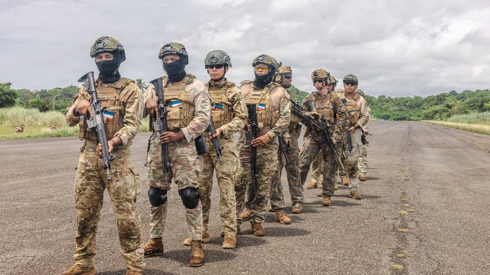 A group of approximately eight to nine armed military or special operations personnel are standing in formation on what appears to be a runway or tarmac in a tropical environment. The soldiers are equipped with tactical gear, body armor, helmets, and assault rifles. Visible flag patches on their uniforms suggest a multinational or joint operation context.