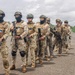 A group of approximately eight to nine armed military or special operations personnel are standing in formation on what appears to be a runway or tarmac in a tropical environment. The soldiers are equipped with tactical gear, body armor, helmets, and assault rifles. Visible flag patches on their uniforms suggest a multinational or joint operation context.