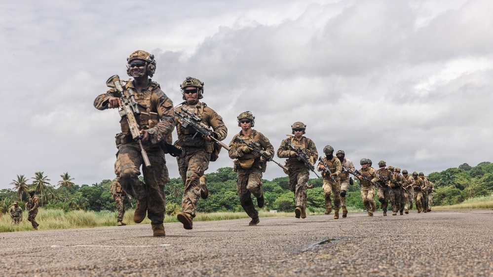 A column of heavily armed military personnel in full combat gear are running along a paved surface, likely a runway or road, in a tropical environment. The soldiers are equipped with rifles, tactical vests, helmets, and eye protection, suggesting a training exercise or tactical operation. The lush green vegetation and palm trees in the background indicate a tropical or subtropical geographic location.