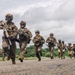 A column of heavily armed military personnel in full combat gear are running along a paved surface, likely a runway or road, in a tropical environment. The soldiers are equipped with rifles, tactical vests, helmets, and eye protection, suggesting a training exercise or tactical operation. The lush green vegetation and palm trees in the background indicate a tropical or subtropical geographic location.