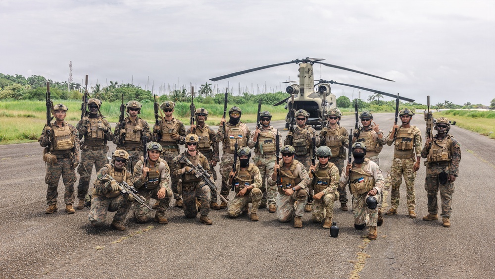 A group of approximately 20 armed military personnel in full combat gear pose for a group photo on a tarmac or airstrip. They are positioned in front of a large tandem-rotor helicopter, identifiable as a CH-47 Chinook. The setting appears to be a tropical or subtropical region based on the surrounding vegetation.