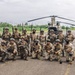 A group of approximately 20 armed military personnel in full combat gear pose for a group photo on a tarmac or airstrip. They are positioned in front of a large tandem-rotor helicopter, identifiable as a CH-47 Chinook. The setting appears to be a tropical or subtropical region based on the surrounding vegetation.