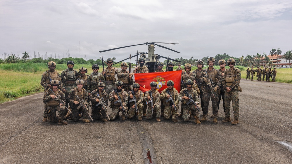 A group of approximately 20-25 armed military personnel pose for a group photograph on a tarmac or airstrip in front of a helicopter. They are holding a red U.S. Marine Corps flag/guidon, suggesting this is a joint or unit photo following a training exercise or operation. The tropical vegetation and architecture in the background indicate a location in a warm, likely Southeast Asian or Latin American environment.