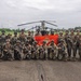 A group of approximately 20-25 armed military personnel pose for a group photograph on a tarmac or airstrip in front of a helicopter. They are holding a red U.S. Marine Corps flag/guidon, suggesting this is a joint or unit photo following a training exercise or operation. The tropical vegetation and architecture in the background indicate a location in a warm, likely Southeast Asian or Latin American environment.