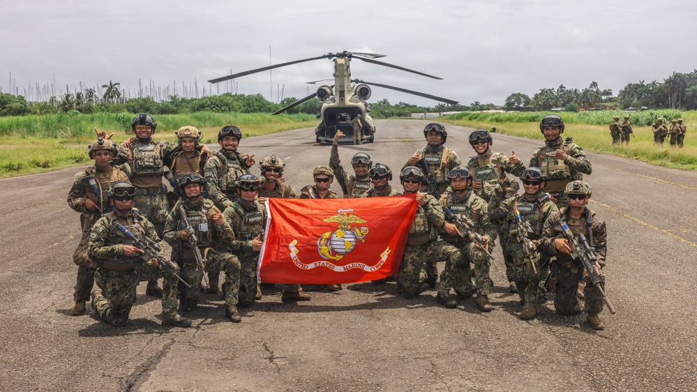 A group of approximately 20 military personnel pose for a group photo on a tarmac or airstrip, holding a red United States Marine Corps flag. Behind them is a CH-47 Chinook tandem-rotor helicopter. Personnel wear a mix of camouflage uniforms, some marked 'MARINA' (indicating Mexican Navy), suggesting a joint U.S.-Mexican military exercise or operation.