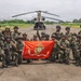 A group of approximately 20 military personnel pose for a group photo on a tarmac or airstrip, holding a red United States Marine Corps flag. Behind them is a CH-47 Chinook tandem-rotor helicopter. Personnel wear a mix of camouflage uniforms, some marked 'MARINA' (indicating Mexican Navy), suggesting a joint U.S.-Mexican military exercise or operation.