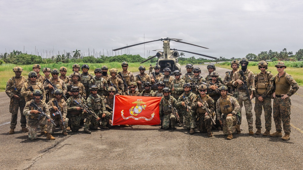 A large group of military personnel pose for a group photograph on a tarmac or airstrip, holding a United States Marine Corps flag. The group appears to include both U.S. Marines and personnel from at least one partner nation, identifiable by different flag patches on their uniforms. A CH-47 Chinook helicopter is visible in the background, suggesting this is a joint military exercise or operation.