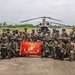 A large group of military personnel pose for a group photograph on a tarmac or airstrip, holding a United States Marine Corps flag. The group appears to include both U.S. Marines and personnel from at least one partner nation, identifiable by different flag patches on their uniforms. A CH-47 Chinook helicopter is visible in the background, suggesting this is a joint military exercise or operation.