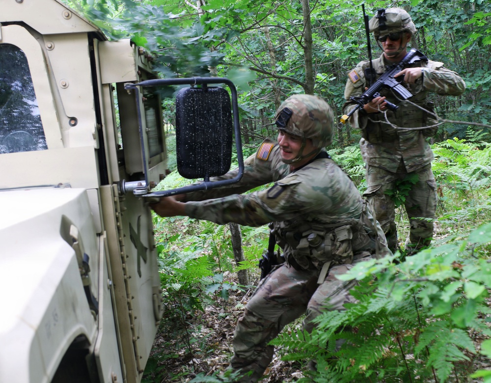 Two U.S. Army soldiers in MultiCam/OCP camouflage uniforms are conducting a tactical exercise in a wooded area alongside a military HMMWV (Humvee). One soldier is opening or securing the vehicle door while crouching, while the second soldier stands in a security posture holding an M4 carbine rifle. Both wear American flag patches on their sleeves, indicating U.S. military affiliation.