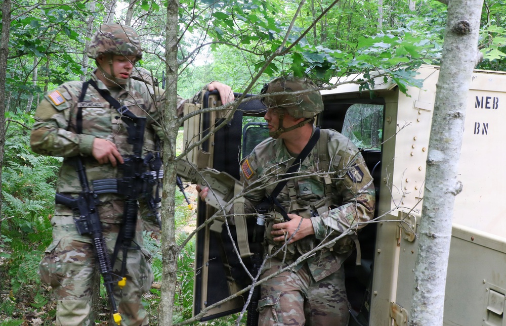 Two U.S. Army soldiers in OCP (Operational Camouflage Pattern) uniforms are conducting what appears to be a field training exercise in a wooded area. One soldier stands alert with an M4-style rifle while the other exits or enters a military HMMWV (Humvee) vehicle. The setting suggests a tactical training scenario in a forested environment.