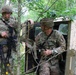 Two U.S. Army soldiers in OCP (Operational Camouflage Pattern) uniforms are conducting what appears to be a field training exercise in a wooded area. One soldier stands alert with an M4-style rifle while the other exits or enters a military HMMWV (Humvee) vehicle. The setting suggests a tactical training scenario in a forested environment.