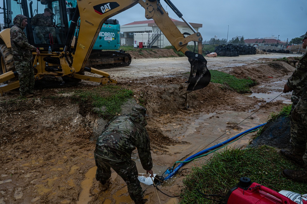 Military personnel in camouflage uniforms are conducting construction or emergency response work in muddy, waterlogged conditions. A yellow CAT excavator and a blue Kobelco excavator are present on the site, with workers managing hoses and equipment in standing water. The overcast, rainy conditions suggest this may be flood mitigation or storm damage response operations.