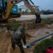 Military personnel in camouflage uniforms are conducting construction or emergency response work in muddy, waterlogged conditions. A yellow CAT excavator and a blue Kobelco excavator are present on the site, with workers managing hoses and equipment in standing water. The overcast, rainy conditions suggest this may be flood mitigation or storm damage response operations.