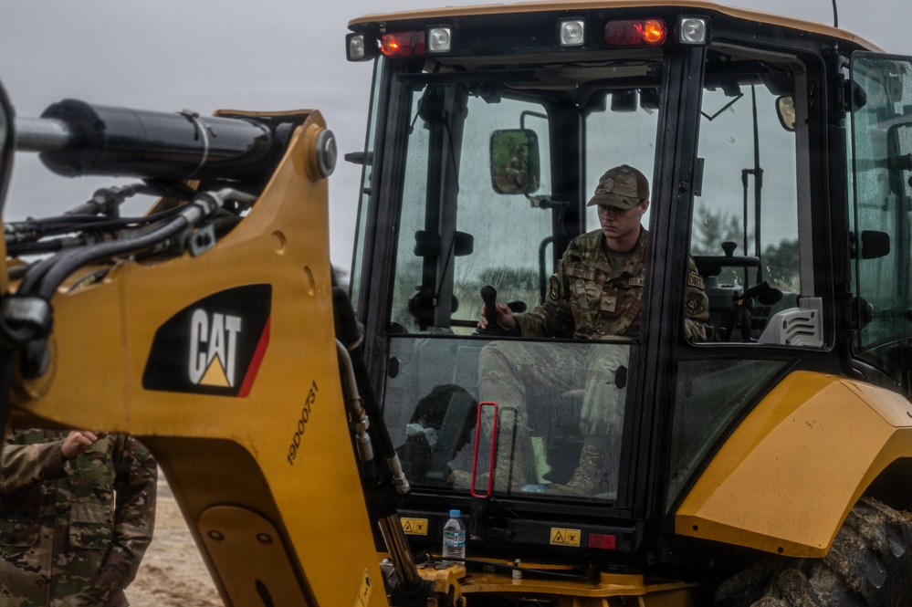 A military service member in camouflage uniform and cap is operating a yellow Caterpillar (CAT) backhoe loader from inside the enclosed cab. The equipment appears to be in use at an outdoor construction or earthmoving site under overcast skies. At least one other uniformed individual is partially visible on the left side of the frame.