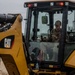 A military service member in camouflage uniform and cap is operating a yellow Caterpillar (CAT) backhoe loader from inside the enclosed cab. The equipment appears to be in use at an outdoor construction or earthmoving site under overcast skies. At least one other uniformed individual is partially visible on the left side of the frame.
