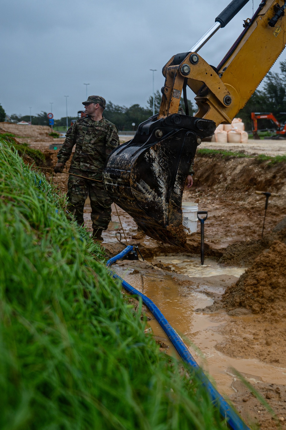 A military service member in camouflage uniform oversees excavation work at what appears to be a construction or utility repair site. A yellow excavator bucket is actively digging in a muddy trench that contains blue pipes and standing water. The overcast conditions and wet soil suggest recent or ongoing rainfall complicating the operation.