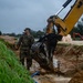 A military service member in camouflage uniform oversees excavation work at what appears to be a construction or utility repair site. A yellow excavator bucket is actively digging in a muddy trench that contains blue pipes and standing water. The overcast conditions and wet soil suggest recent or ongoing rainfall complicating the operation.