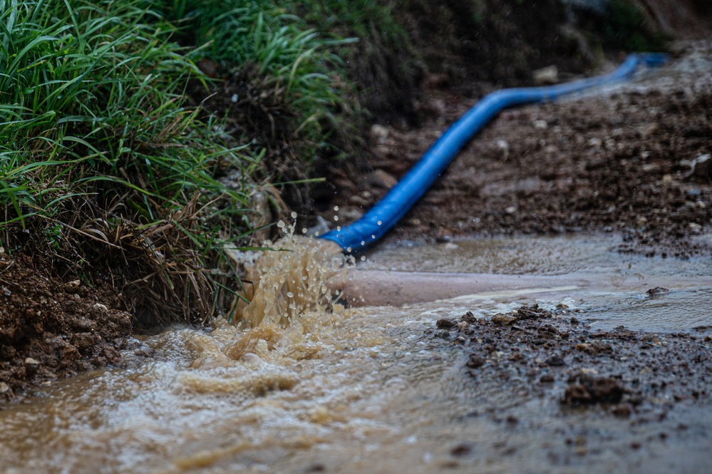 A blue hose or pipe is actively discharging a large volume of muddy, sediment-laden water onto a dirt surface near a grassy embankment. The water is splashing forcefully, creating turbulence and pooling in the surrounding muddy terrain. This appears to be a dewatering or pumping operation, likely at a construction or excavation site.