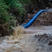 A blue hose or pipe is actively discharging a large volume of muddy, sediment-laden water onto a dirt surface near a grassy embankment. The water is splashing forcefully, creating turbulence and pooling in the surrounding muddy terrain. This appears to be a dewatering or pumping operation, likely at a construction or excavation site.